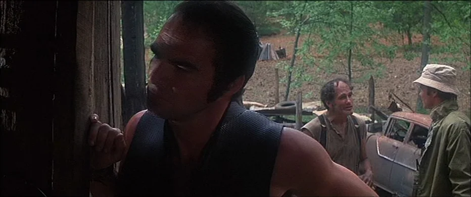 Deliverance (1972) movie still: medium — An open wooden structure overlooks a rural clearing containing a rusted vintage car. Three men stand in the…