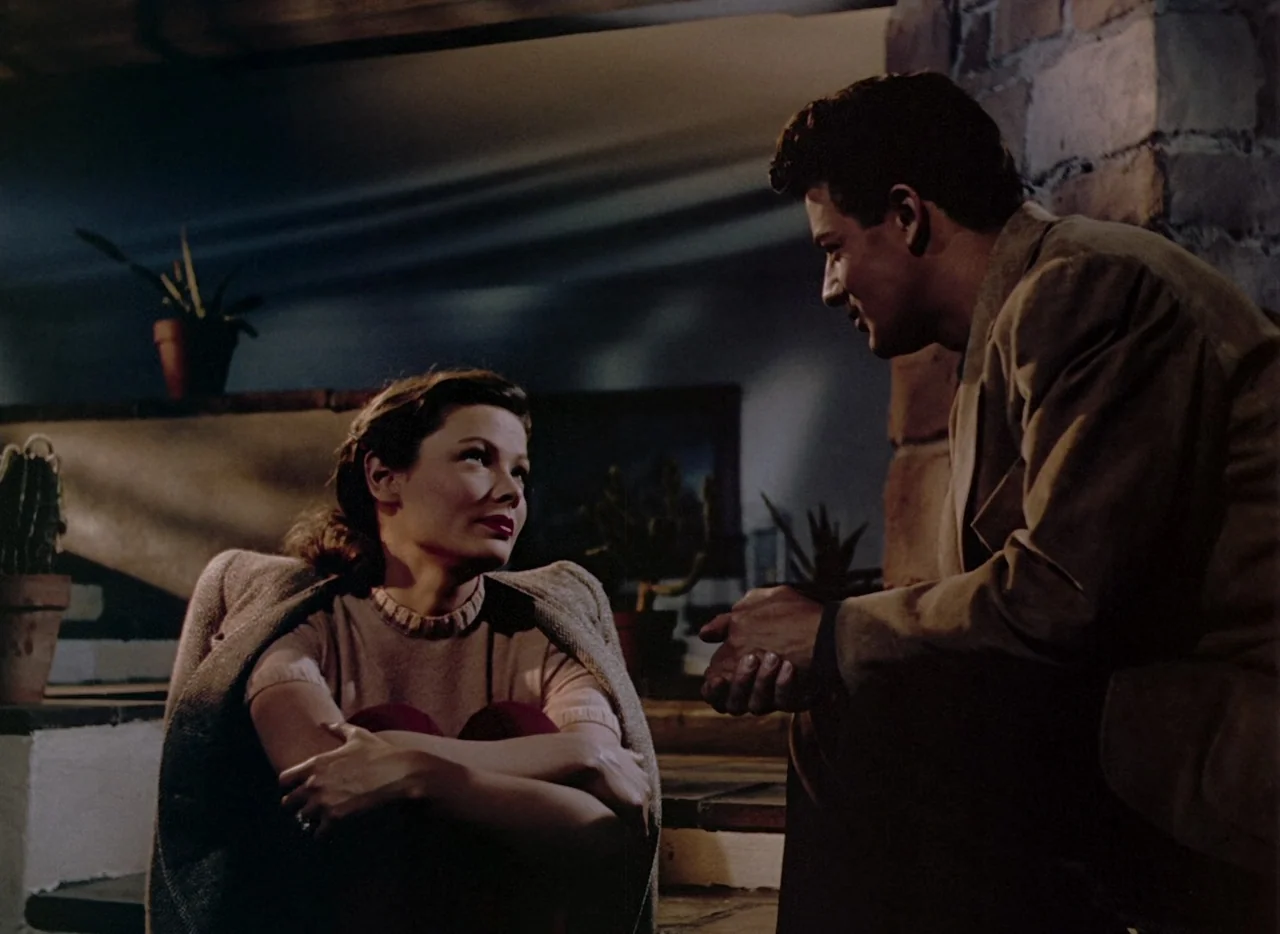 Leave Her To Heaven (1945) movie still: medium — A dimly lit room with brick accents and potted plants arranged in the background. A woman sits on…