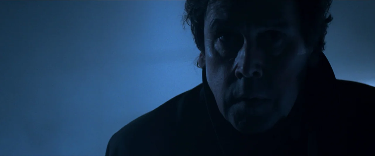 V for Vendetta (2006) movie still: closeup — A dimly lit interior space is bathed in a uniform blue light. A man wearing a dark coat looks forward…