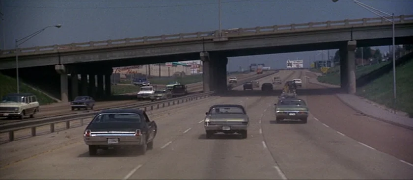 Nashville (1975) movie still: wide — A wide concrete highway stretches under a large overpass with multiple vintage vehicles traveling. Cars move in…