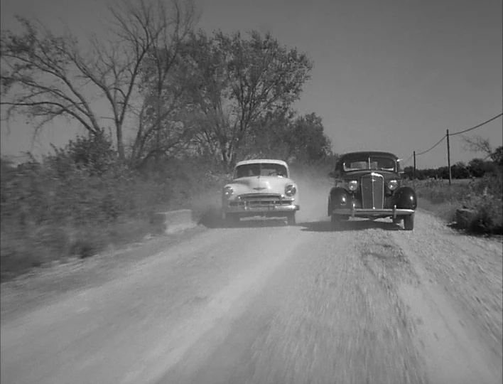 Carnival of Souls (1962) movie still: wide — A dusty dirt road winds through sparse, leafless trees under a clear sky. Two vintage mid-century cars…