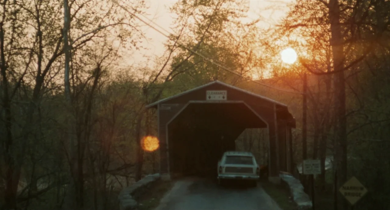 The Rain People (1969) movie still: wide — A wooden covered bridge sits amidst sparse trees during the late afternoon. A vintage station wagon is…
