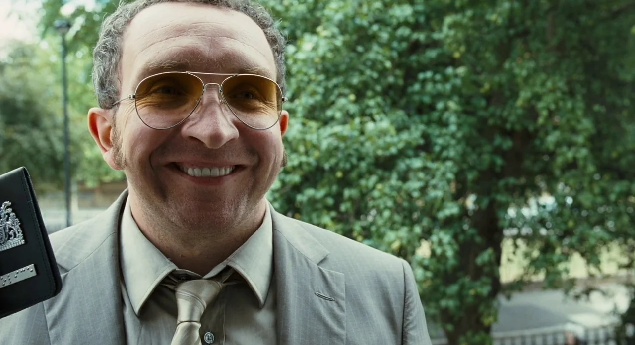 London Boulevard (2010) movie still: closeup — A man stands outdoors in front of out-of-focus green trees and shrubs. He grins widely while…