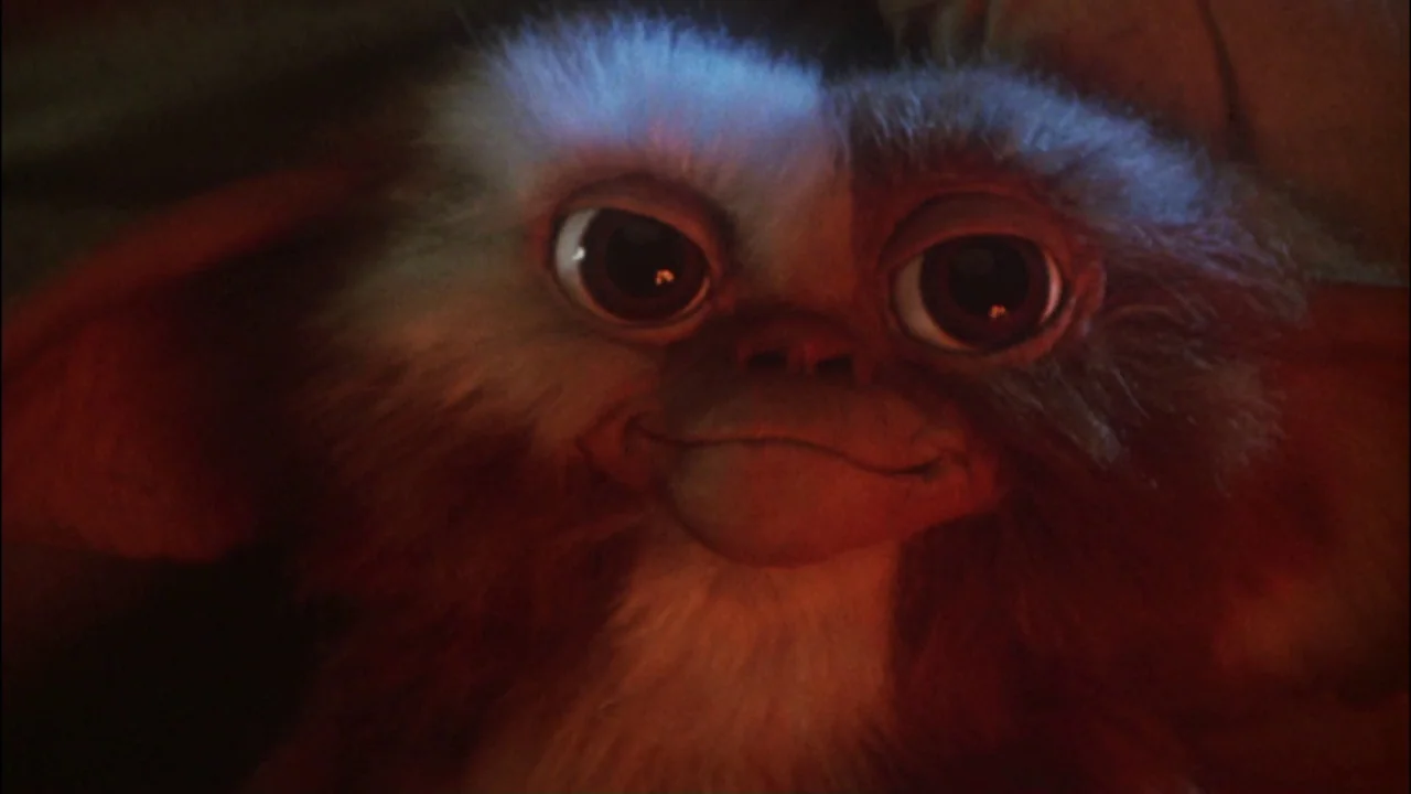 Gremlins (1984) movie still: closeup — A dimly lit room provides the backdrop for a close-up of a small creature. The creature faces the camera…