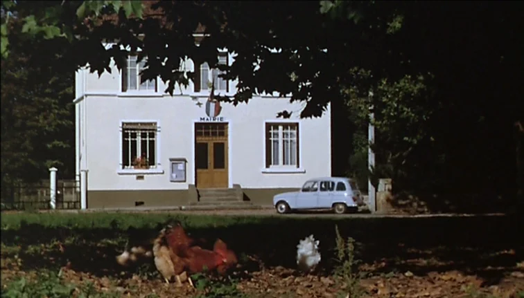 Le Boucher (1970) movie still: wide — A rural French administrative building with white stucco walls stands under large leafy trees. Chickens forage…