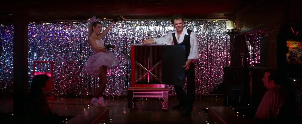Boogie Nights movie still: wide — A dimly lit stage features a shimmering pink tinsel backdrop behind a magic box. A woman in a sparkly dress and a…