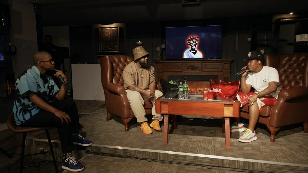 African Throne World Tour (2024) movie still: medium — Three men sit in leather armchairs in a dark studio setting with a television display. They…