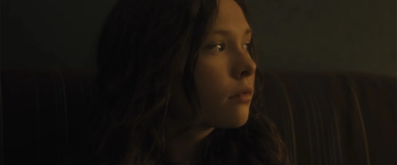 Skin (2009) movie still: closeup — A dark, dimly lit room contains a young woman sitting in the shadows. The subject gazes off-camera toward an…