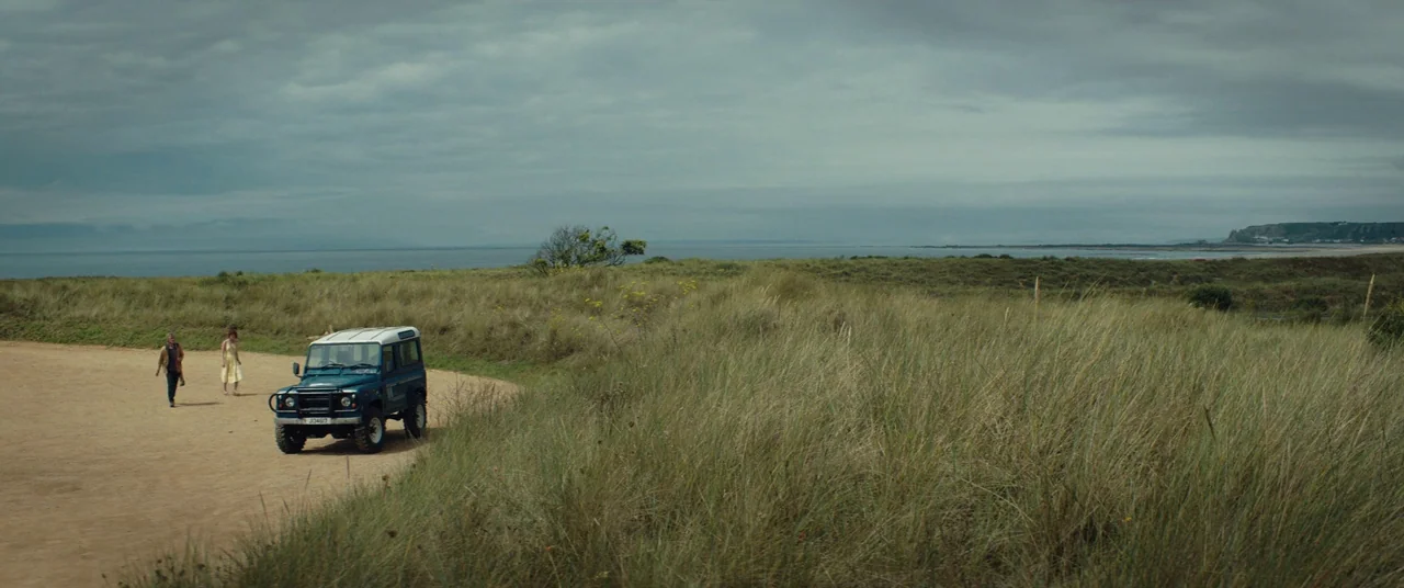 Beast (2017) movie still: wide — An overcast coastal landscape features rolling sand dunes covered in tall, dry grass. A blue Land Rover Defender is…