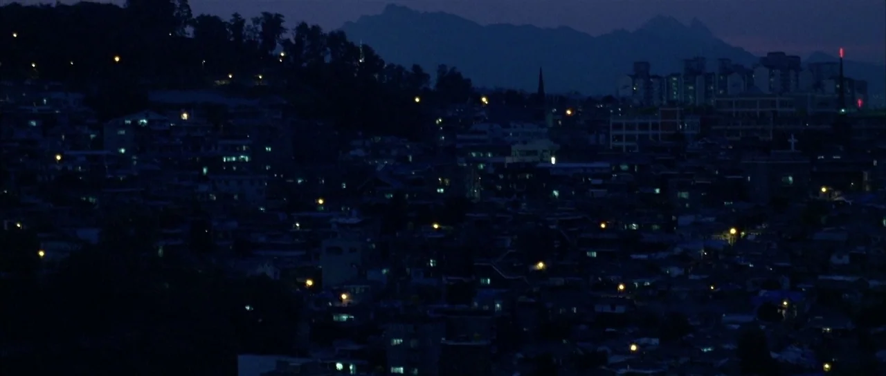 Sympathy for Mr. Vengeance (2002) movie still: establishing — A dense urban hillside at night is dotted with small, glowing artificial lights. Faint…