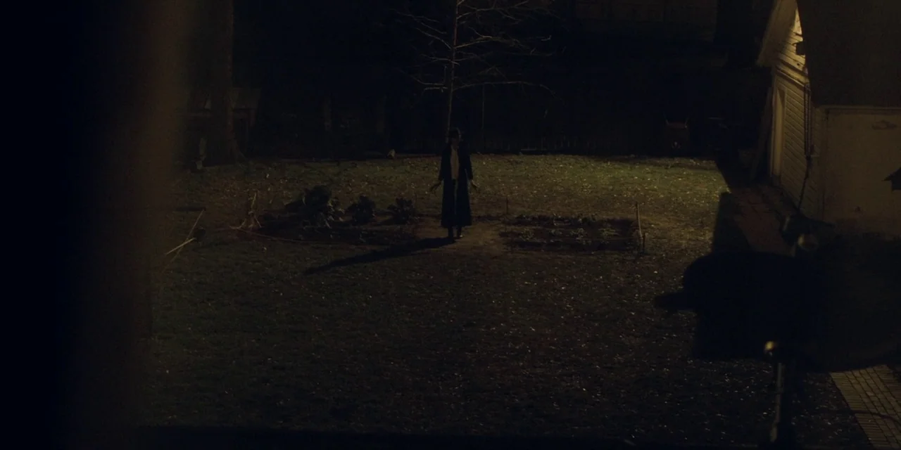 Jakob’s Wife (2021) movie still: wide — A dark, grassy residential backyard at night features a small vegetable plot in center. A woman stands alone…