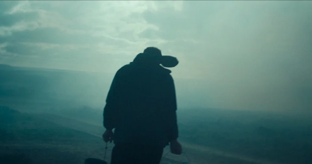God’s Own Country (2017) movie still: medium — A figure walks through a vast field engulfed in thick morning mist. The person carries buckets while…