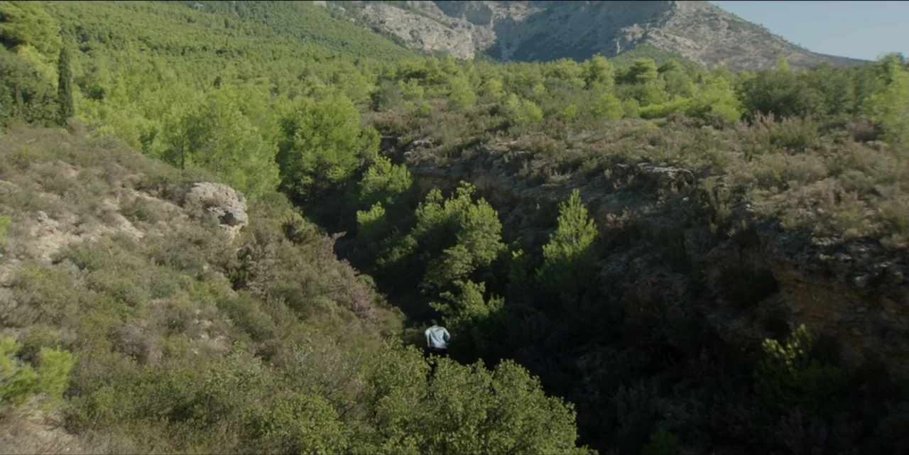 The Hunted (2024) movie still: wide — A deep rocky ravine covered with green pine trees and low shrubs. A person in a light blue shirt walks through…