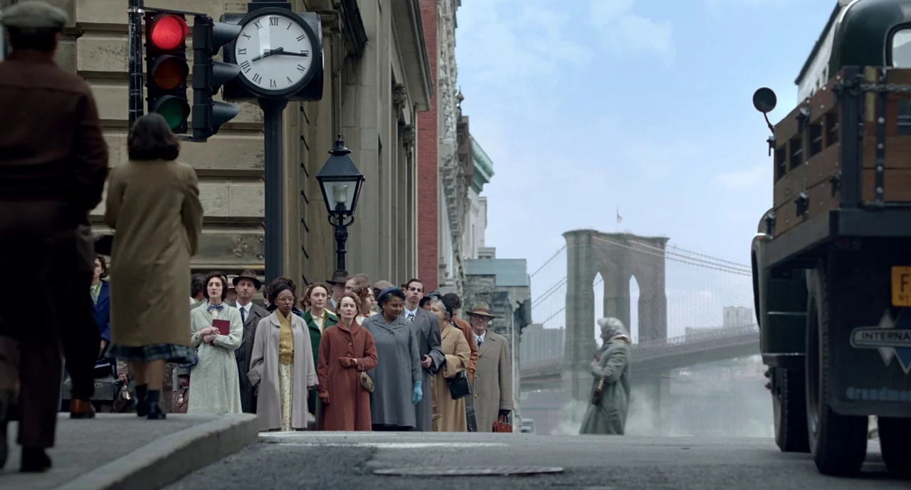 Brooklyn (2015) movie still: wide — A 1950s New York city street features a crowd waiting near a traffic light. The Brooklyn Bridge is visible in…