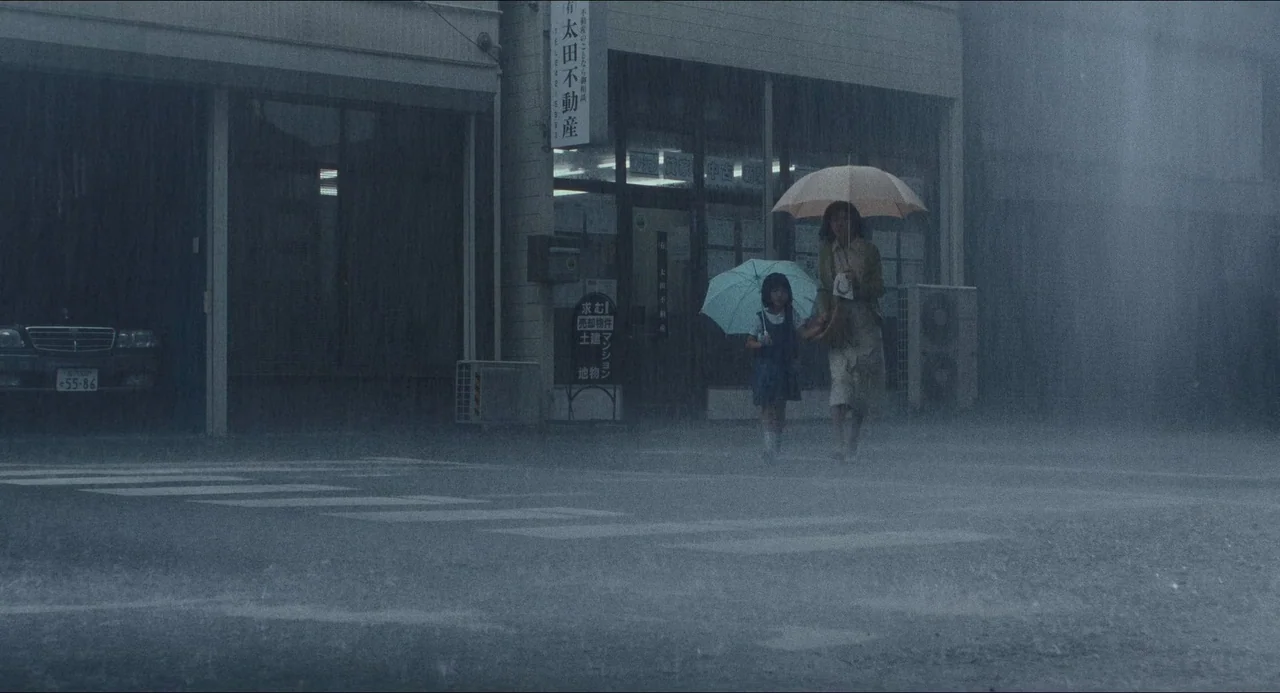 Dark Water (2002) movie still: wide — A rain-drenched street in Japan sits before a storefront with Japanese signage. A woman holds a large umbrella…