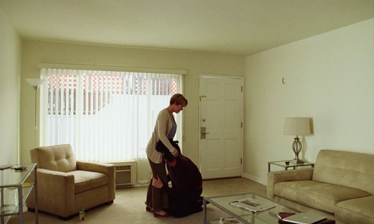 Marriage Story movie still: wide — A sparse, neutral-toned living room features a beige sofa, armchair, and a glass coffee table. A woman stands in…