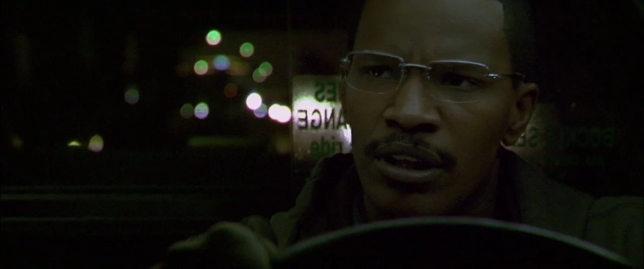 Collateral (2004) movie still: closeup — A dark car interior at night with a city landscape out of focus outside. A man in glasses grips the…