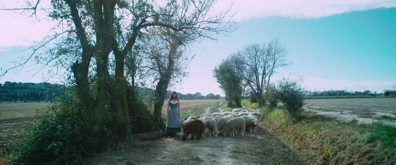 At Eternity’s Gate (2018) movie still: wide — A dirt path runs through a wide agricultural field flanked by sparse deciduous trees. A young woman in…