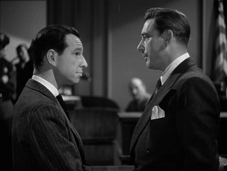 The Postman Always Rings Twice (1946) movie still: medium — A courtroom interior features a wooden bench and a blurred figure in the background. Two…