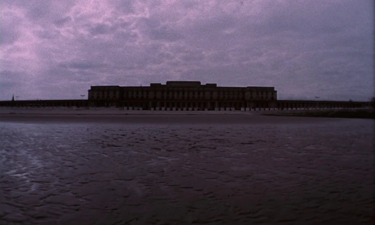 Daughters of Darkness (1971) movie still: establishing — A long, multi-story building sits on the sand behind a stretch of dark water. The sky is…