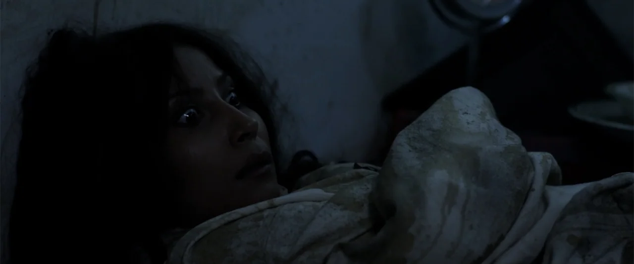 Mermaid Down (2019) movie still: closeup — A dimly lit room with a person partially visible under a stained blanket. A young woman lies in bed with…