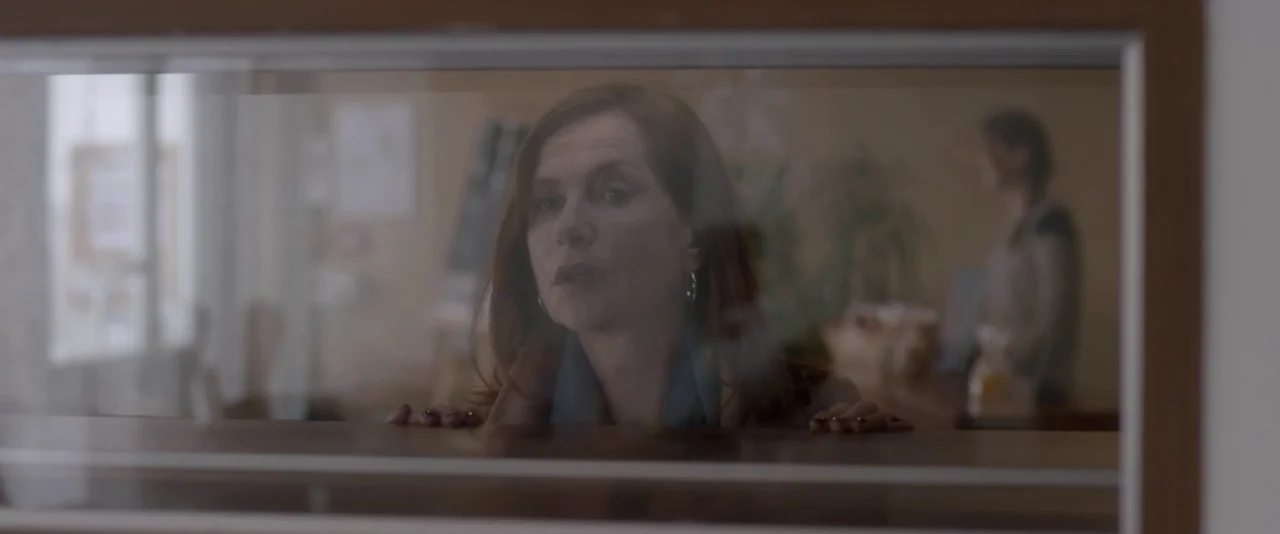 Elle (2016) movie still: closeup — A woman leans over a wooden counter seen through a transparent glass partition. In the blurred background,…