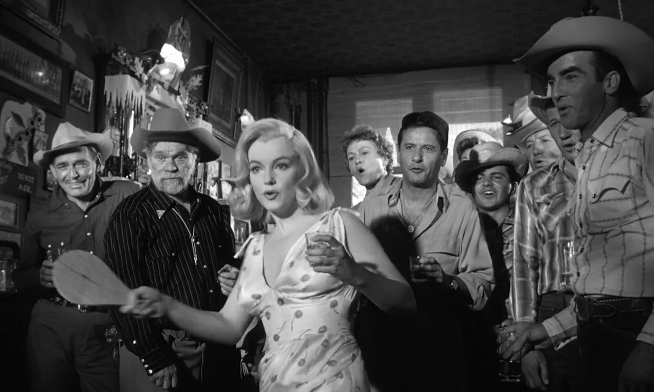 The Misfits (1961) movie still: medium — A crowded bar interior with framed pictures and dark walls lining the background. A group of men and one…