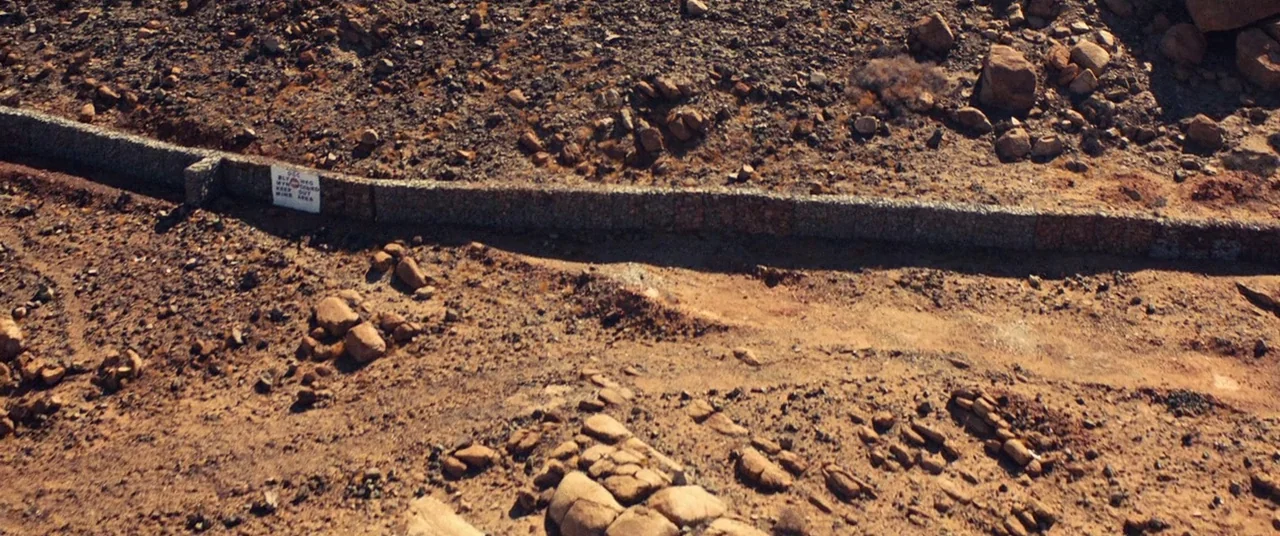 Uncut Gems (2019) movie still: establishing — A rugged desert landscape features rocky terrain and a long, low gabion retaining wall. A small white…