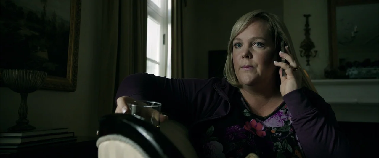 K.G.F: Chapter 2 (2022) movie still: medium — A dimly lit room with draped windows and dark furniture. A woman sits on a sofa, holding a smartphone…