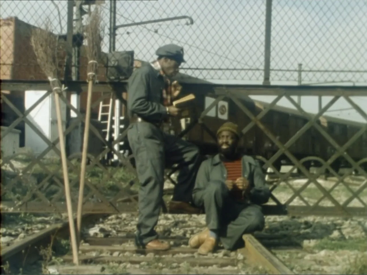 A Dessert for Constance (1981) movie still: fullbody — Two men rest on gravel tracks next to a chain-link fence by rail cars. The standing man reads…