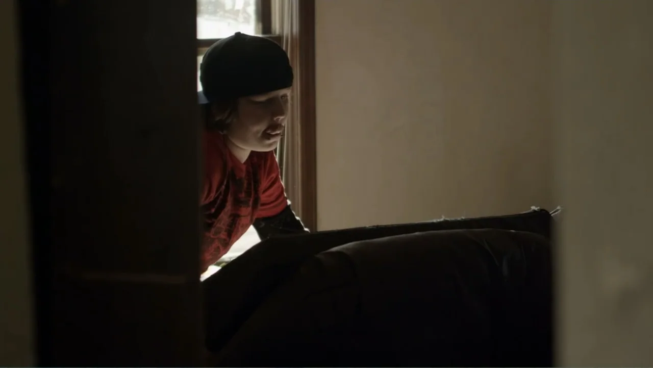 Rich Hill (2014) movie still: medium — A residential room features a doorway partition and a dark leather couch. A teenager in a red shirt peers…