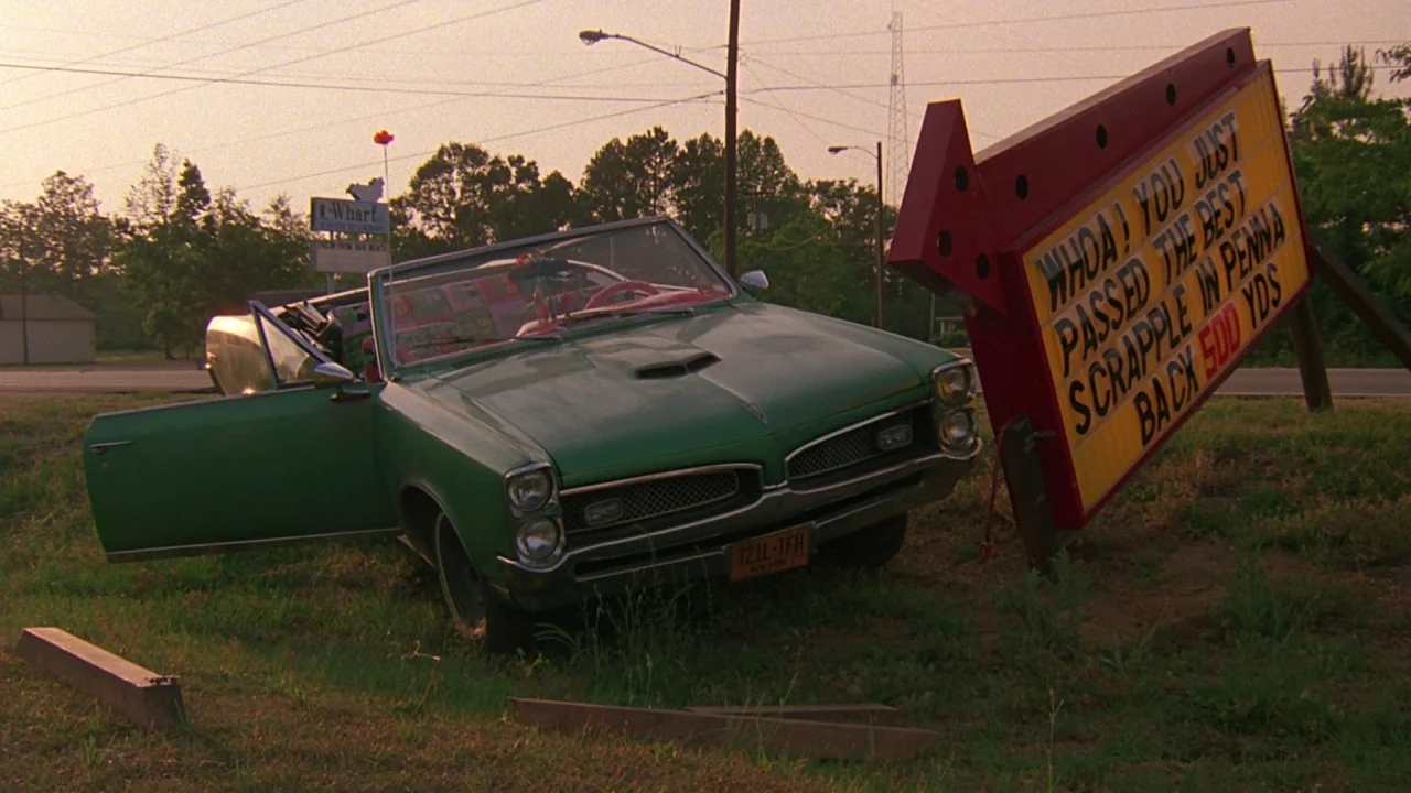 Something Wild (1986) movie still: wide — A rural roadside grassy area features a green convertible crashed into a large sign. The sign features…