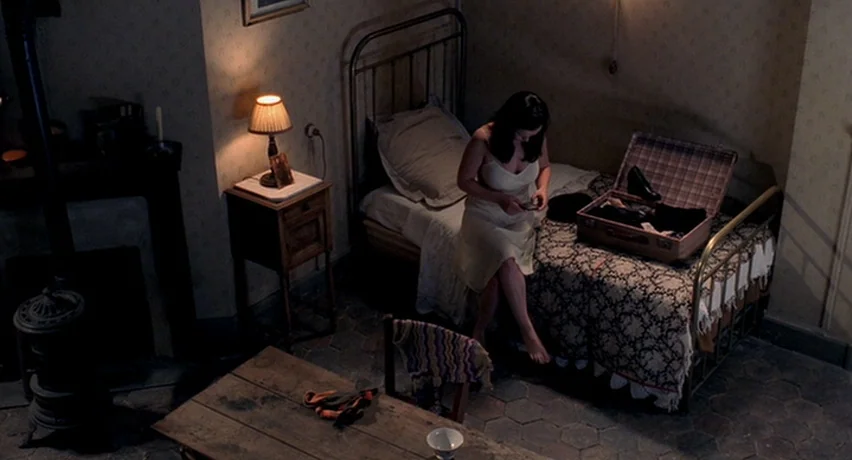 The Man Who Cried (2000) movie still: wide — A sparsely furnished room features a metal frame bed and a wooden nightstand. A woman sits on the edge…