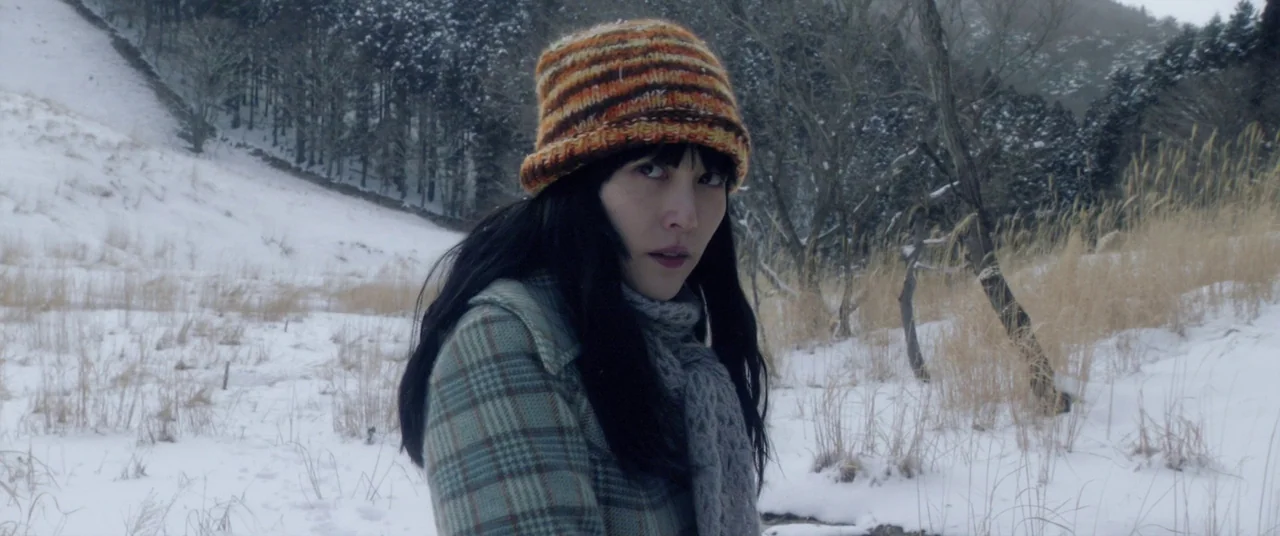 Norwegian Wood (2010) movie still: medium — A snow-covered field sits before a dense, dark forest of bare winter trees. A young woman in a plaid…