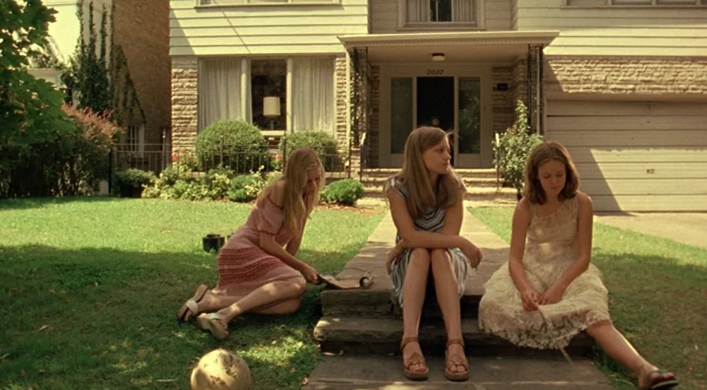 The Virgin Suicides (1999) movie still: wide — A suburban lawn with a stone walkway leading to a two-story residential home. Three teenage girls are…