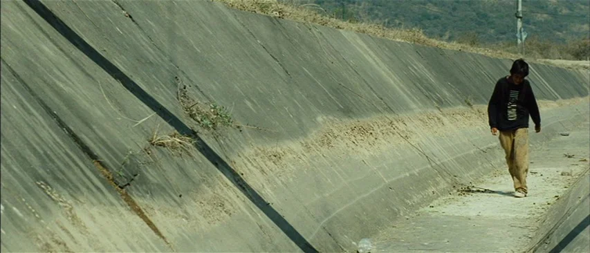 The Headless Woman (2008) movie still: wide — A dry, slanted concrete drainage canal cuts diagonally through the frame under bright sunlight. A lone…