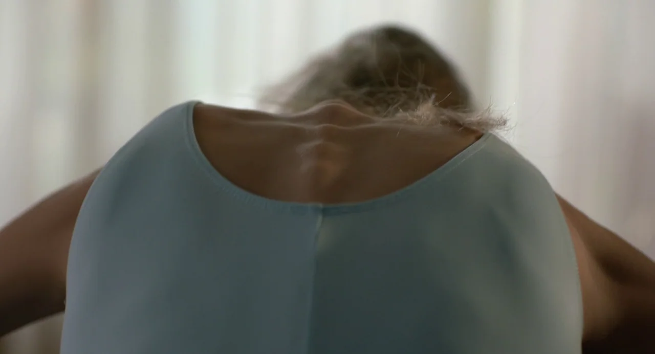Hatching (2022) movie still: closeup — A minimalist interior space with white curtains blurred in the background. A woman in a pale blue top bends…