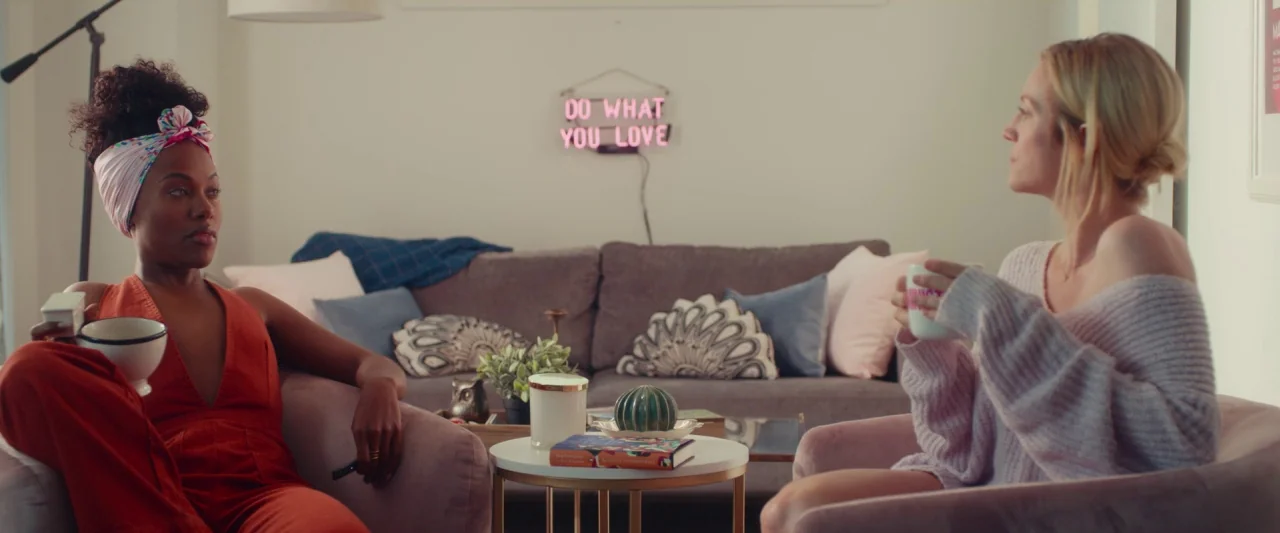 Someone Great (2019) movie still: medium — A well-lit room contains a gray sofa adorned with pillows and decorative objects. Two women sit in…
