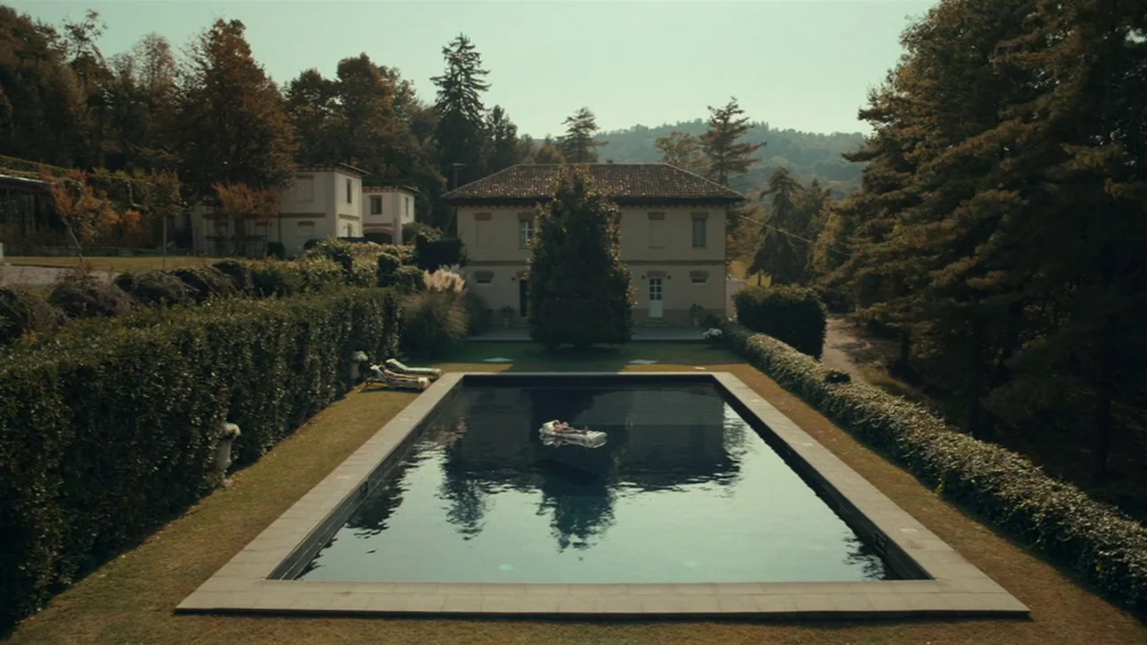 Amanda (2022) movie still: wide — A rectangular swimming pool sits in front of a tan two-story Italian villa. A lone person rests on a white…