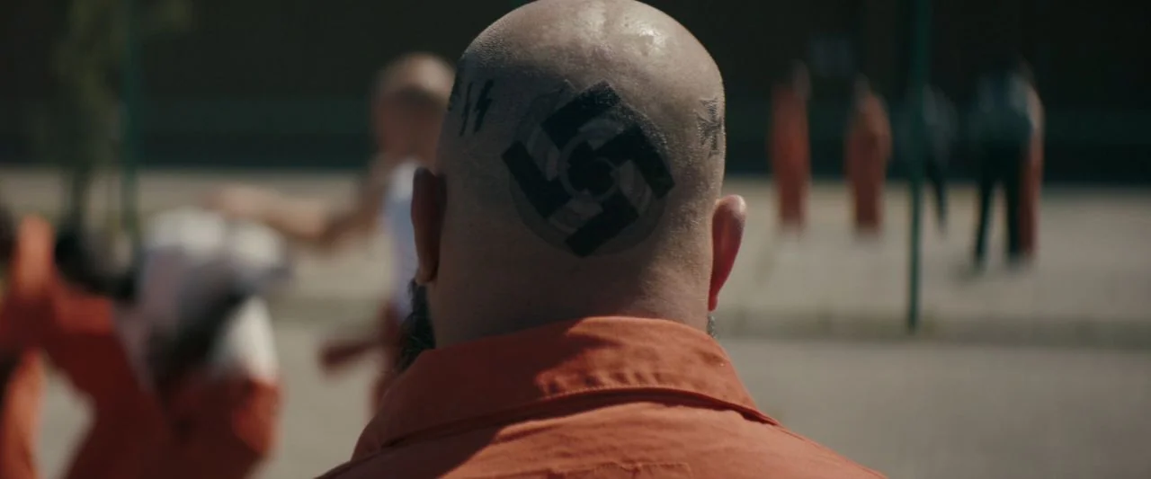 Becky (2020) movie still: closeup — An outdoor prison yard features blurred figures in orange uniforms moving in the background. A man stands in the…
