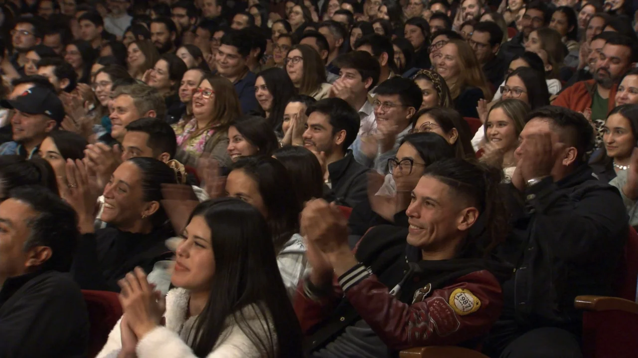 Felipe Avello: Doglover (2024) movie still: medium — A crowded auditorium filled with people sitting in red upholstered theater seats. A large…