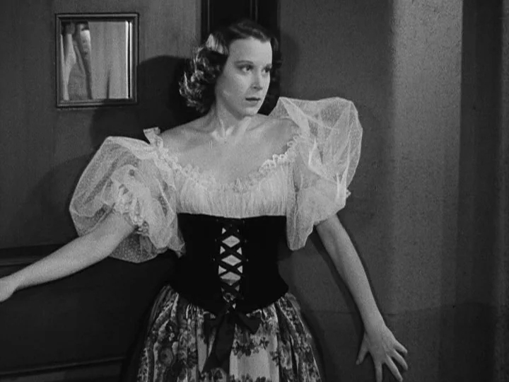 A Night at the Opera (1935) movie still: medium — A woman stands in a dimly lit hallway beside a door with a window. She wears an elaborate period…