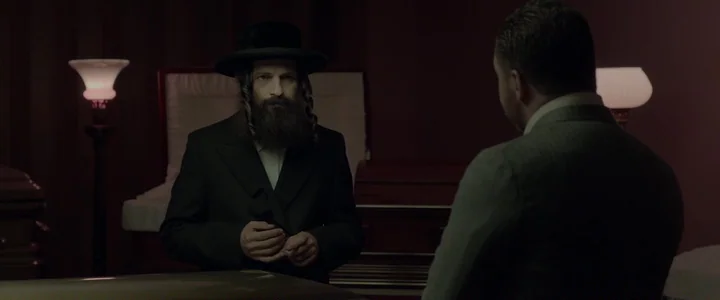 To Dust - Nightmares (2018) movie still: medium — A dark funeral parlor with two floor lamps flanking a wooden casket. A man with payot stands…