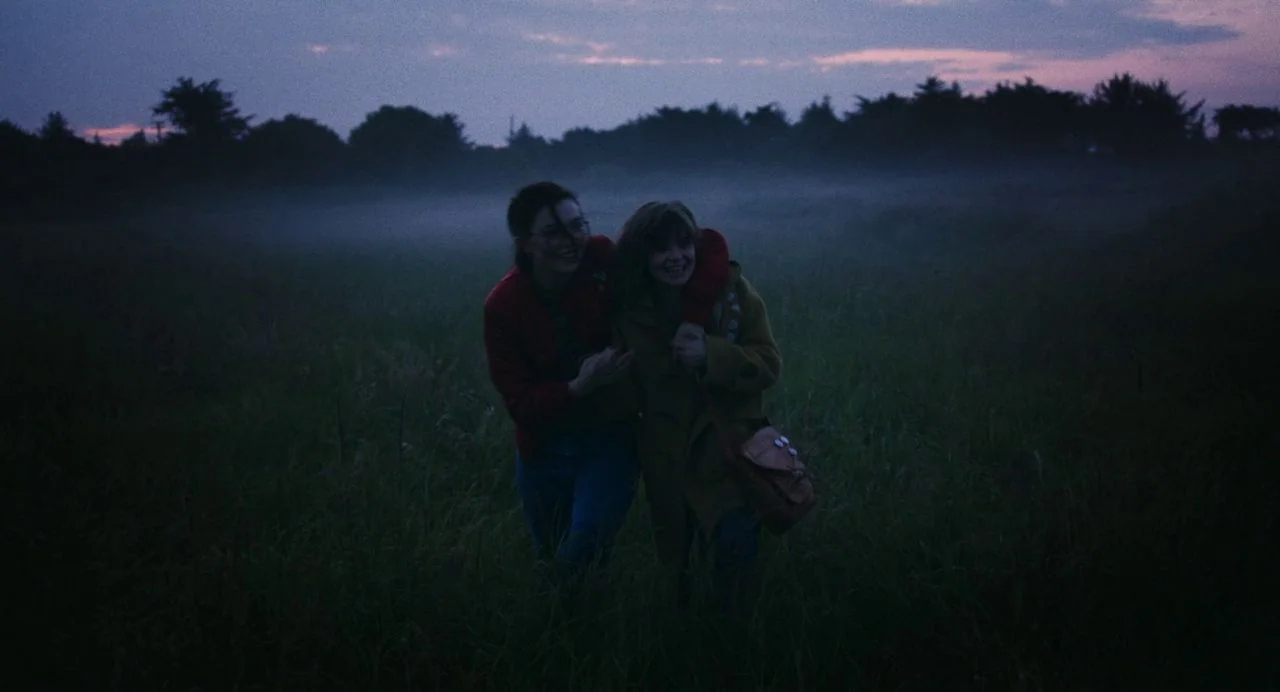 Dating Amber movie still: fullbody — Two women walk through a foggy, dark grass field at dusk. The sky has a faint purple and blue glow behind the…