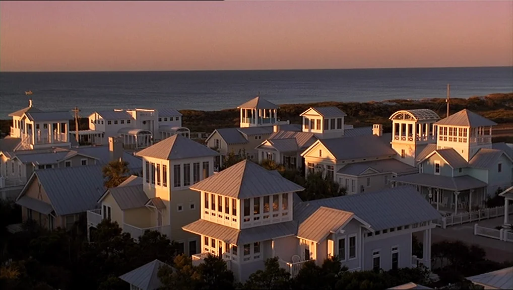 The Truman Show movie still: establishing — A seaside neighborhood of white and pastel houses with metal roofs overlooks the ocean. Palm trees and…