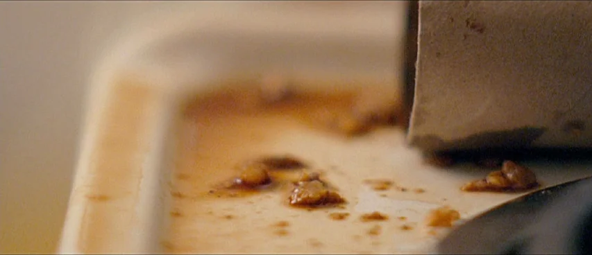 This Movie Is Broken (2010) movie still: closeup — An off-white plate contains scattered remnants of brown sauce and solid food scraps. A cardboard…