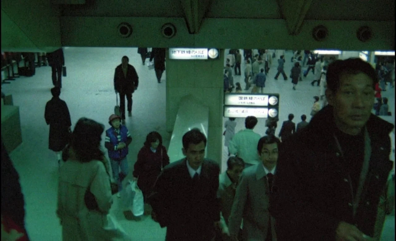 Sans Soleil (1983) movie still: wide — A crowded Japanese subway station concourse with concrete pillars and directional signage. Commuters walk in…