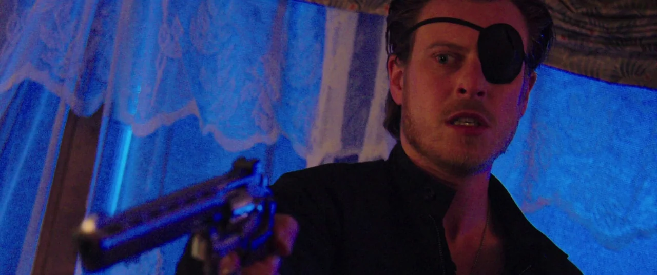 The Mind’s Eye (2015) movie still: medium — A man in a dark jacket stands before a window covered by translucent curtains. He holds a revolver aimed…