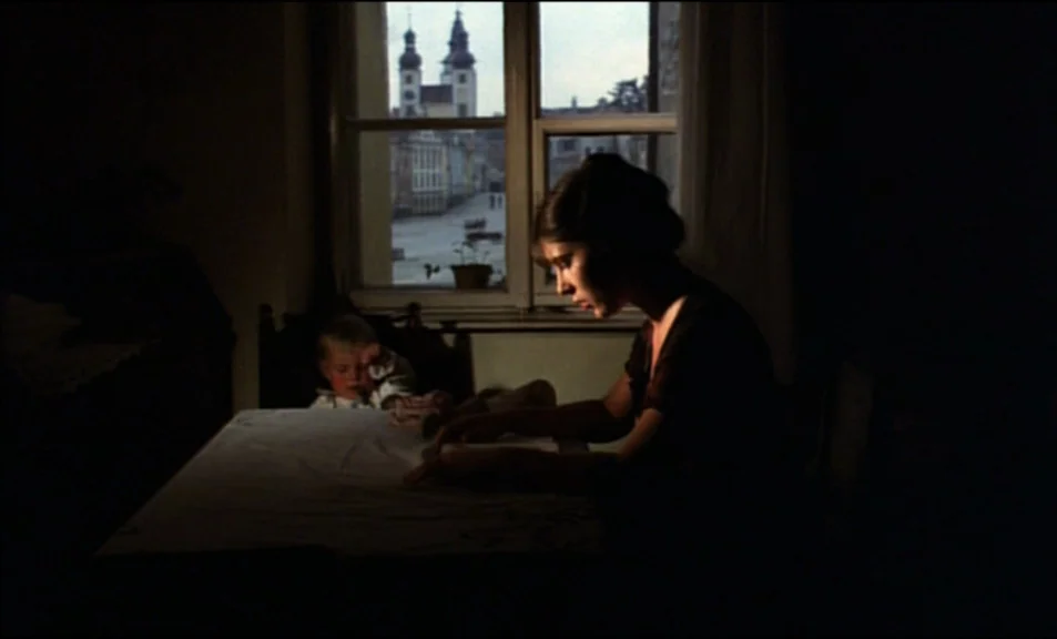 Woyzeck (1979) movie still: medium — A dimly lit room features a woman at a table by a bright window. Through the window, a town square and stone…