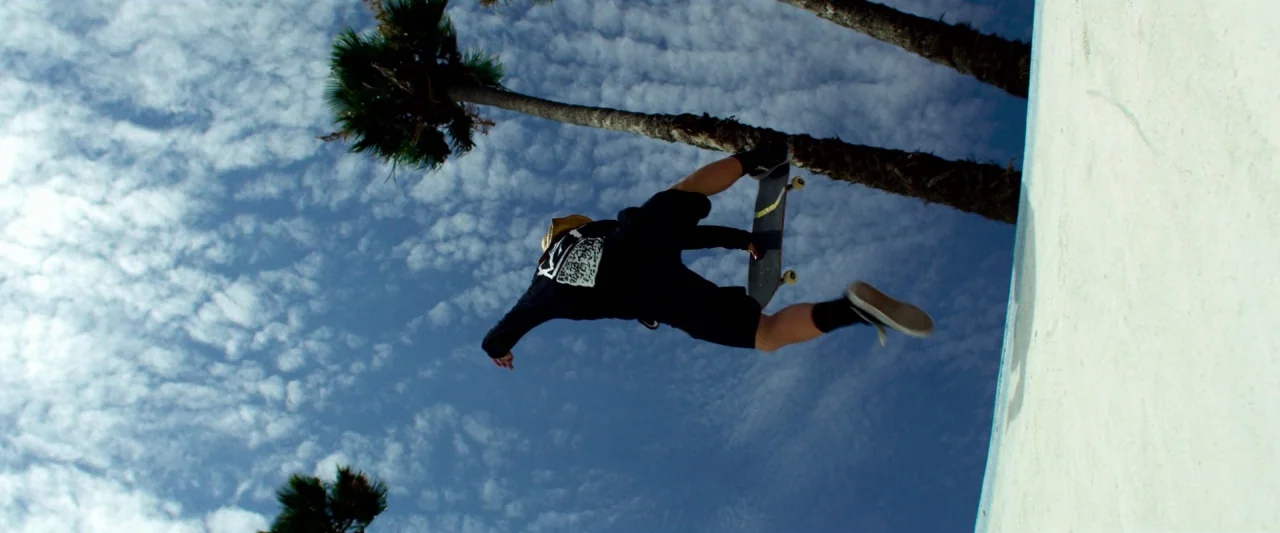 6 Underground (2019) movie still: wide — An outdoor concrete skatepark bowl sits under a bright blue sky with scattered clouds. A skateboarder jumps…